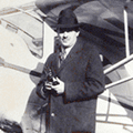 A man in a hat and coat standing next to an airplane.